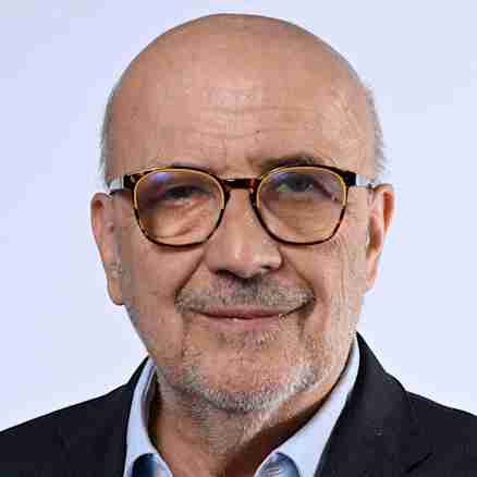 A bald man with glasses, a beard, and a mustache stands in front of a plain background. He is wearing a dark suit with a light blue button-up shirt. His hands are casually placed in his pockets, and he smiles at the camera.