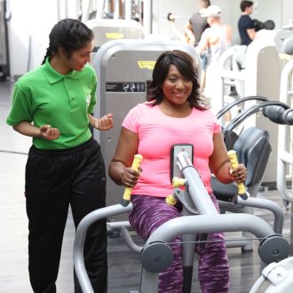 Image shows a female gym instructor helping a female gym member on a piece of resistance gy equipment to help build strength in the arms. 