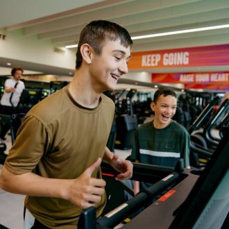 image shows a teenager running on a treadmill with another teenager beside him both are smiling