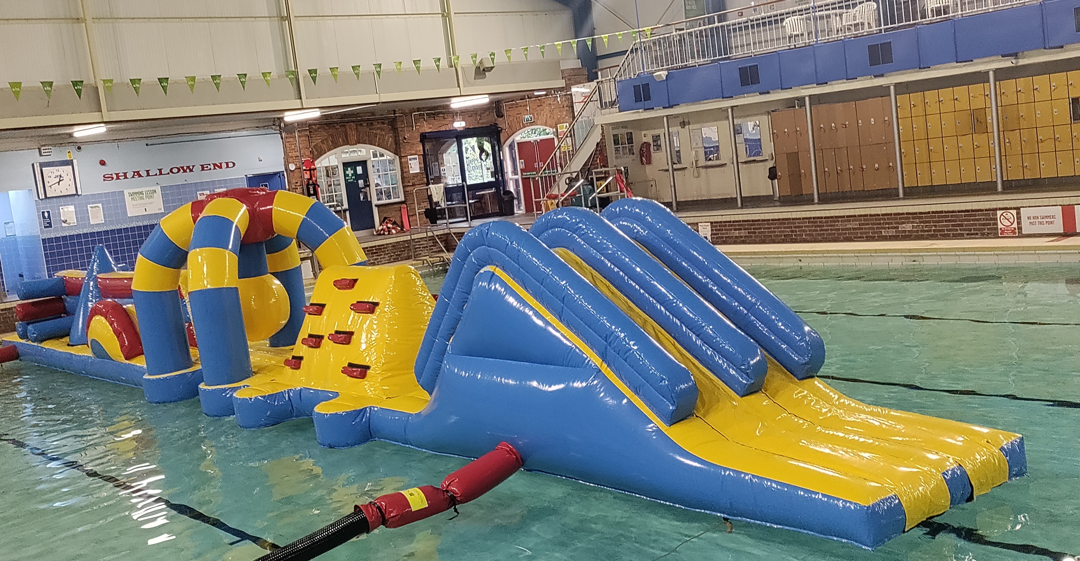 Inflatable water obstacle course in a pool, featuring slides, climbing wall, and arches. It's colourful, mainly yellow and blue, with a fun, playful vibe.