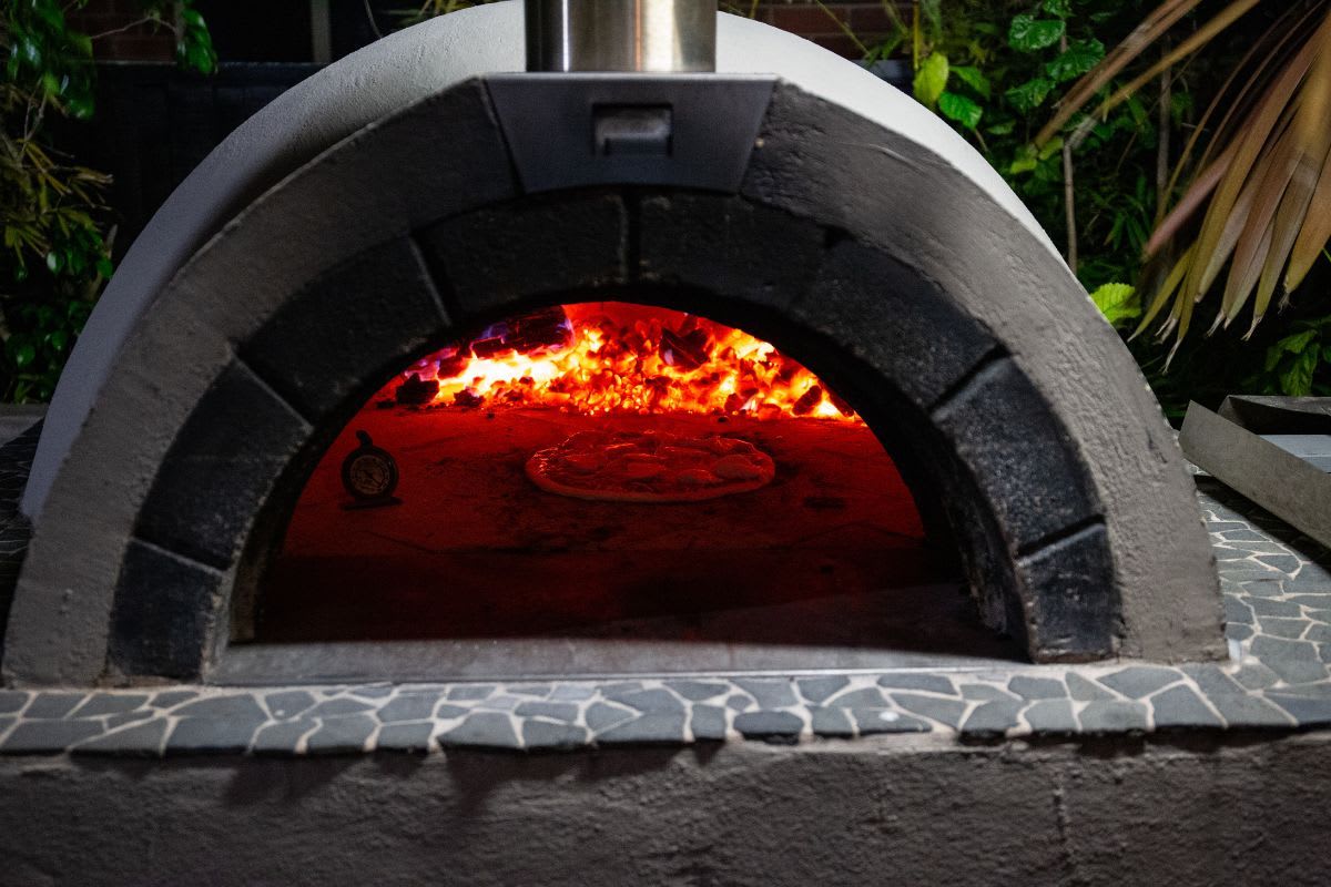 Ein befeuerter Pizzaofen im Garten mit einer offenen Flammenfront.
