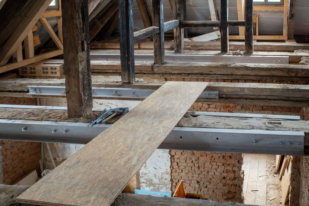Renovierungsarbeiten in einem älteren Gebäude mit sichtbaren Stahlträgern und einem Holzbrett über einem Träger, umgeben von traditionellem Holzgebälk und Mauerwerk.