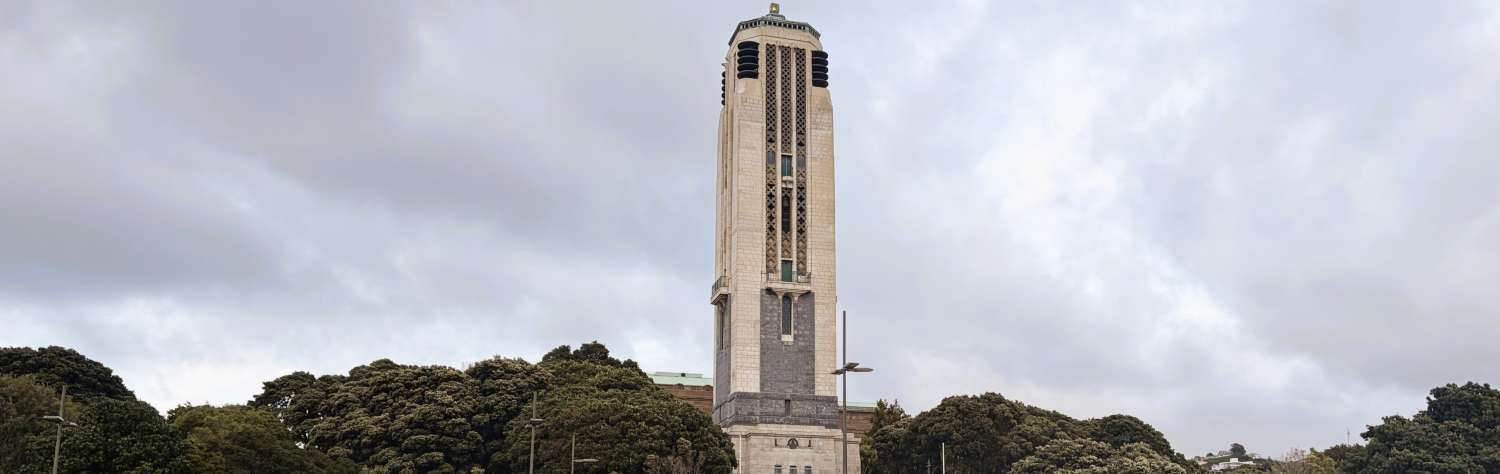 Tall stone clock tower standing behind a plaza and surrounded by green trees under a cloudy sky.