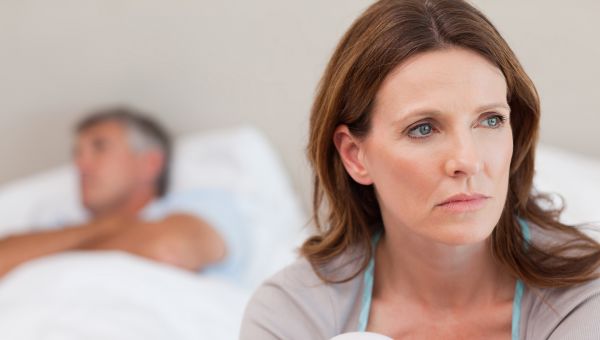 woman sitting up in bed looking sad, with sleeping husband in background