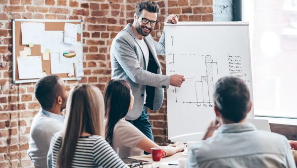 Handsome young man in glasses standing near whiteboard and pointing on the chart while his coworkers listening and sitting at the table