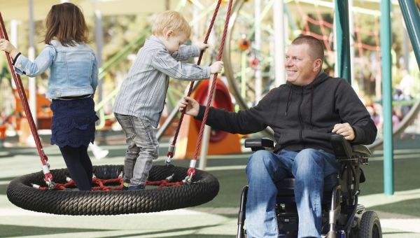 man in a wheelchair looking after two kids playing in playground