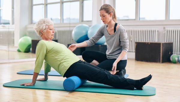 Physical therapist working with active senior woman at rehab. Old woman exercising using foam roller with personal trainer at gym.