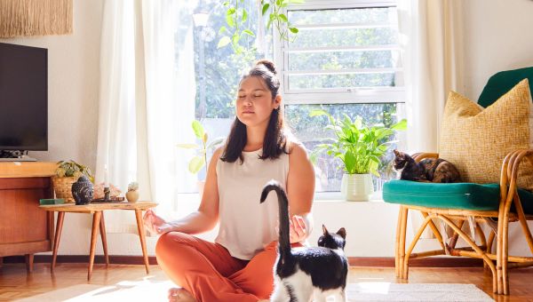 Young person sitting on a yoga mat at home, with a black and white cat in the foreground