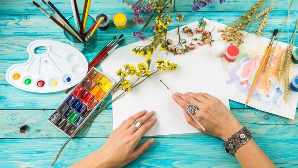 Hand holding a paint brush surrounded by flowers and paints.