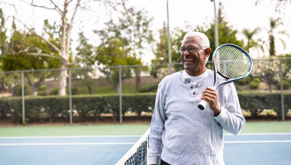 an older fit Black man stands on a tennis court holding a tennis racket and ball 