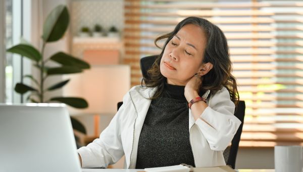 a middle aged Asian woman with pain in her neck rests her neck against her hand while she works at her desk