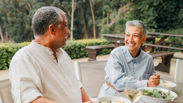 an older Latina woman with short gray hair and an older Latino man enjoy eating healthy food, including salad, in an outdoor restaurant patio