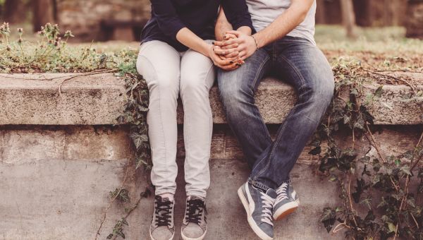a young heterosexual couple sits on a wall holding hands, we cannot see their faces