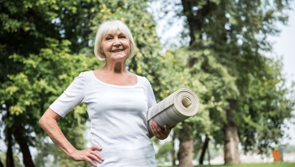 an older woman looking fit and happy smiles as she holds a yoga mat