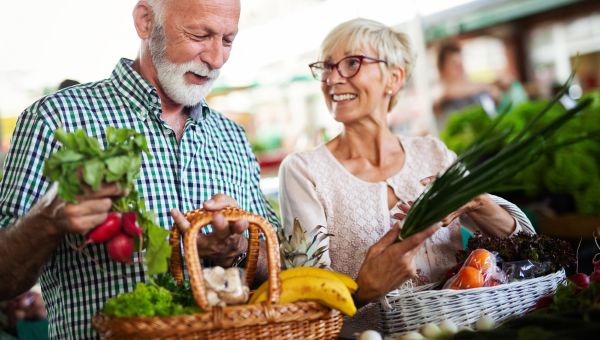 an older white man and woman fill their basket with fresh fruits and vegetables at a farmers' market