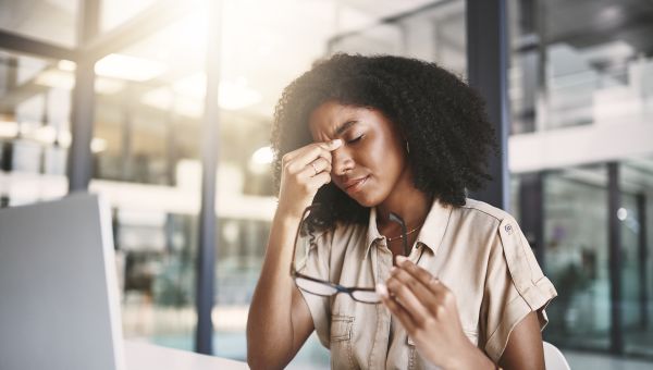 a middle aged Black professional woman sits at her computer, holding her brow as a result of a migraine headache