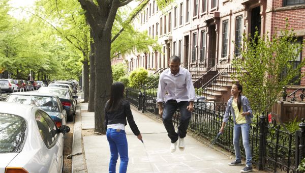 a young black man in a professional outfit jumps rope "Double Dutch" with two young girls on a city residential sidewalk