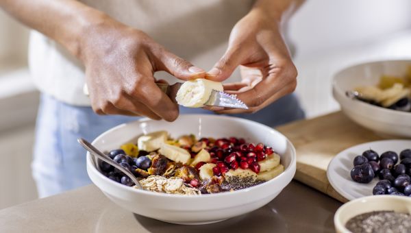 closeup of a Black woman's hands as she prepares a healthy meal of sliced fruit, granola, seeds, and berries