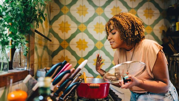 a Black woman stands in a colorfully decorated kitchen, smelling the contents of a pot of healthy stew cooking on her oven range top