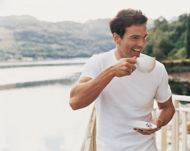 Man Standing Laughing as he Drinks From a Coffee Cup on a Balcony Beside a Lake