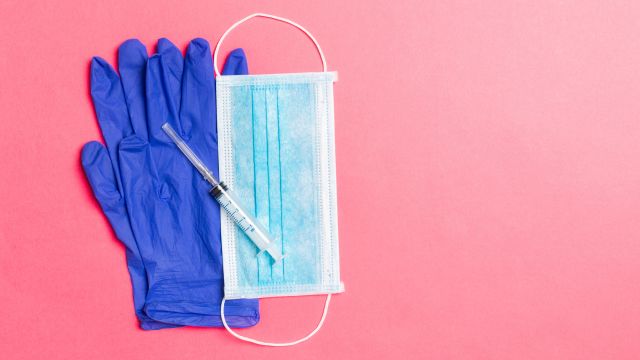A mask, syringe, and gloves rest on a pink background. The shingles vaccine is given as a shot to the upper arm and can be administered by a pharmacist.