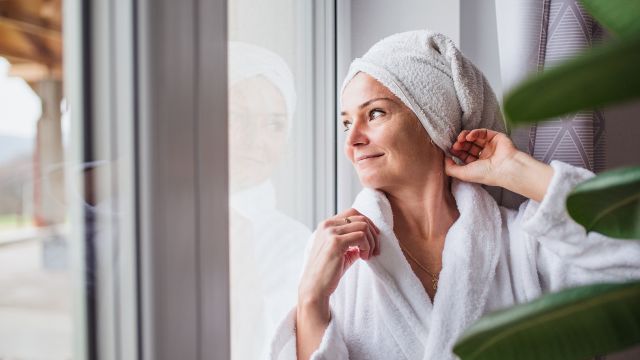 woman in a bath robe with towel-wrapped hair looking out a window