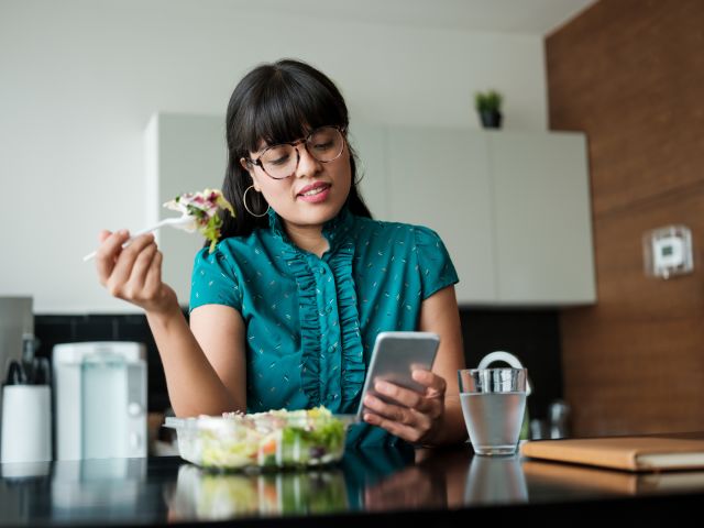 a young woman eats a healthy salad while checking her smartphone
