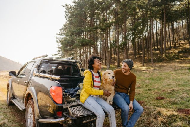 A happy couple on vacation sits in the bed of a pickup truck with a shaggy dog.