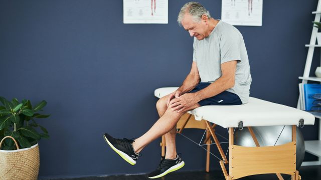 An older man with silver hair experiences knee pain while waiting in the examination room at the doctor's office.