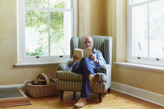 an older white man sits in a sun-lit room, reading a book and boosting his brain health in the process