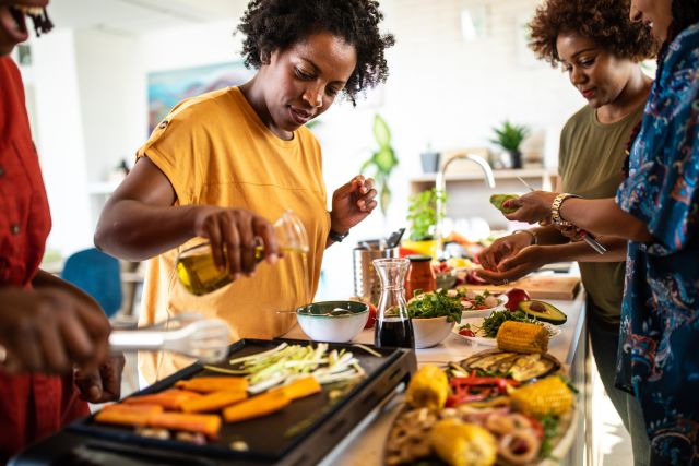 Black woman pouring olive oil onto vegetables that are being cooked on a grill, while surrounded by friends or family who are also cooking