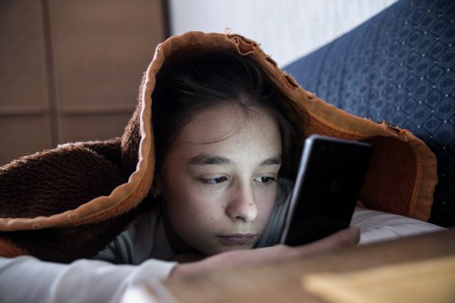 An unhappy teenage girl looks at a smartphone screen while under her covers in bed. 