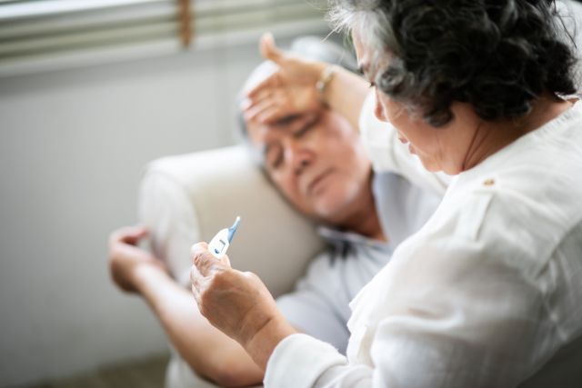 Older person sick with the flu lying on sofa while a caretaker is holding and looking at a thermometer