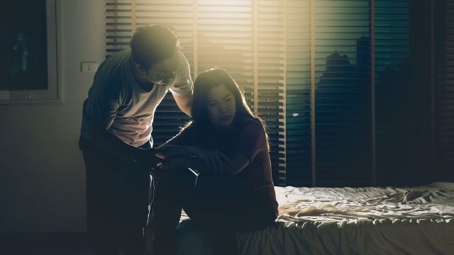 Man consoles sad woman in dark bedroom.