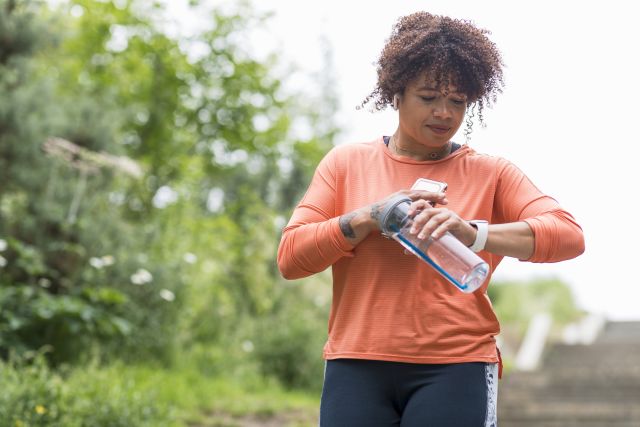 An adult woman walking outdoors, she is holding a water bottle and her smartphone, she is checking the time on her smartwatch.