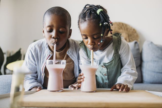 Young brother and sister sitting on their living room sofa, leaning over the coffee table to drinks smoothies from a straw