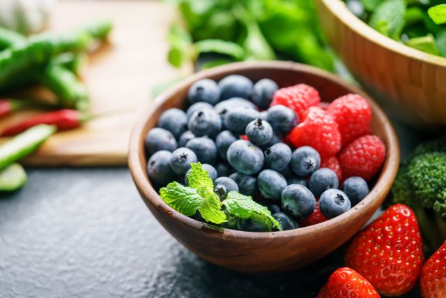 closeup of a bowl of blueberries and raspberries, surrounded by green vegetables