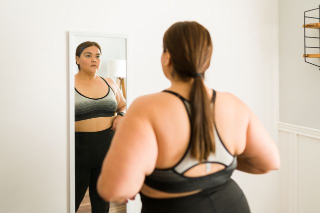 full figured woman looks worriedly in a full length mirror