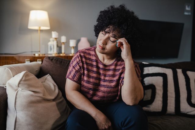 Fatigued woman sitting on her couch, eyes closed with head in hand