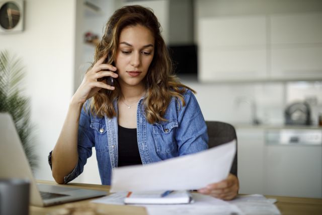 Woman sits at kitchen table looking over paperwork while she talks to her insurance company on the phone 