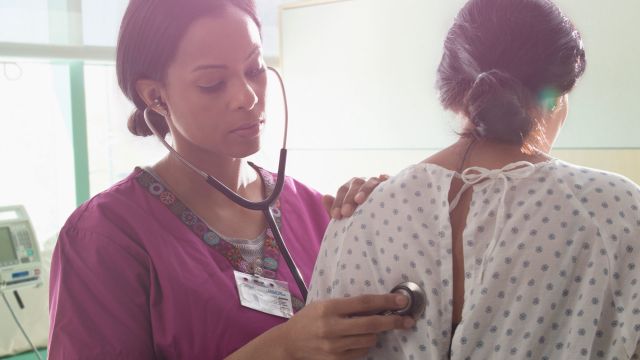 A young Asian man is examined by a White doctor, who is listening to the patient's lungs with a stethoscope.