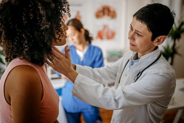 doctor examining a woman's neck for thyroid enlargement in an exam room, nurse in the background