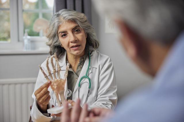 Mature Female Doctor Meeting With Male Patient Discussing Arthritis Pain In Hand Using Anatomical Model