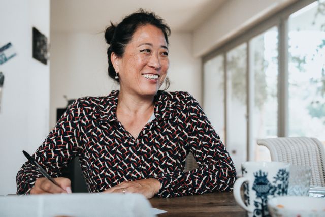 A woman living with fibromyalgia smiles at her dining room table while she journals in a diary about her symptoms.