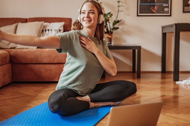 A woman meditates and stretches in her living room. Stress can contribute to atopic dermatitis symptoms, and it's recommended to find ways to reduce stress.