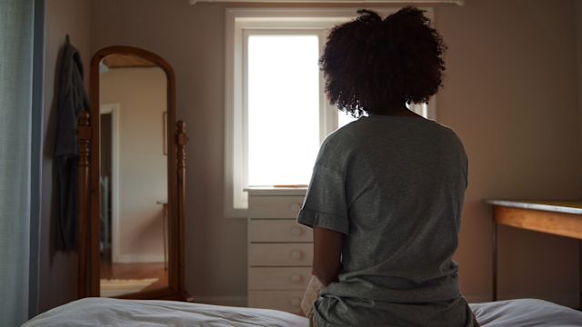 A young woman with psoriasis sits in her bedroom during the daytime, struggling with low mood and motivation.