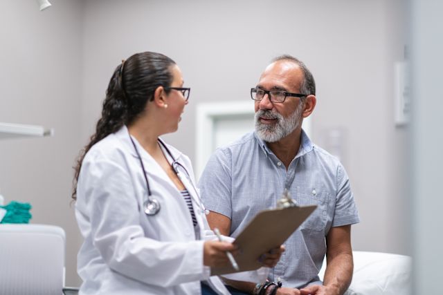 a middle aged female doctor with glasses and a long black braid speaks with her patient, an older white man with glasses and a beard, about steatotic liver disease, also known as fatty liver disease  