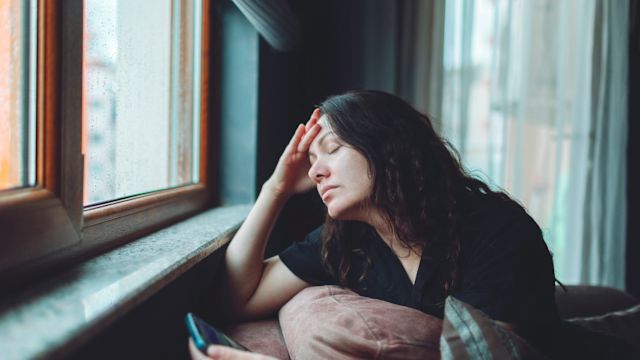 A depressed woman sits alone at home scrolling through her phone while looking out a window. Graves' disease can affect moods, behavior, and cognition, and also cause structural changes to the brain.