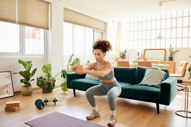 A young, fit Black woman in workout clothing performs a squat in her living room as she exercises at home