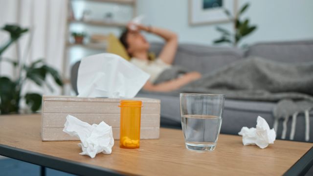 A woman is home sick with the flu, resting on the couch. A glass of water, medicine, and tissues are on the coffee table nearby.